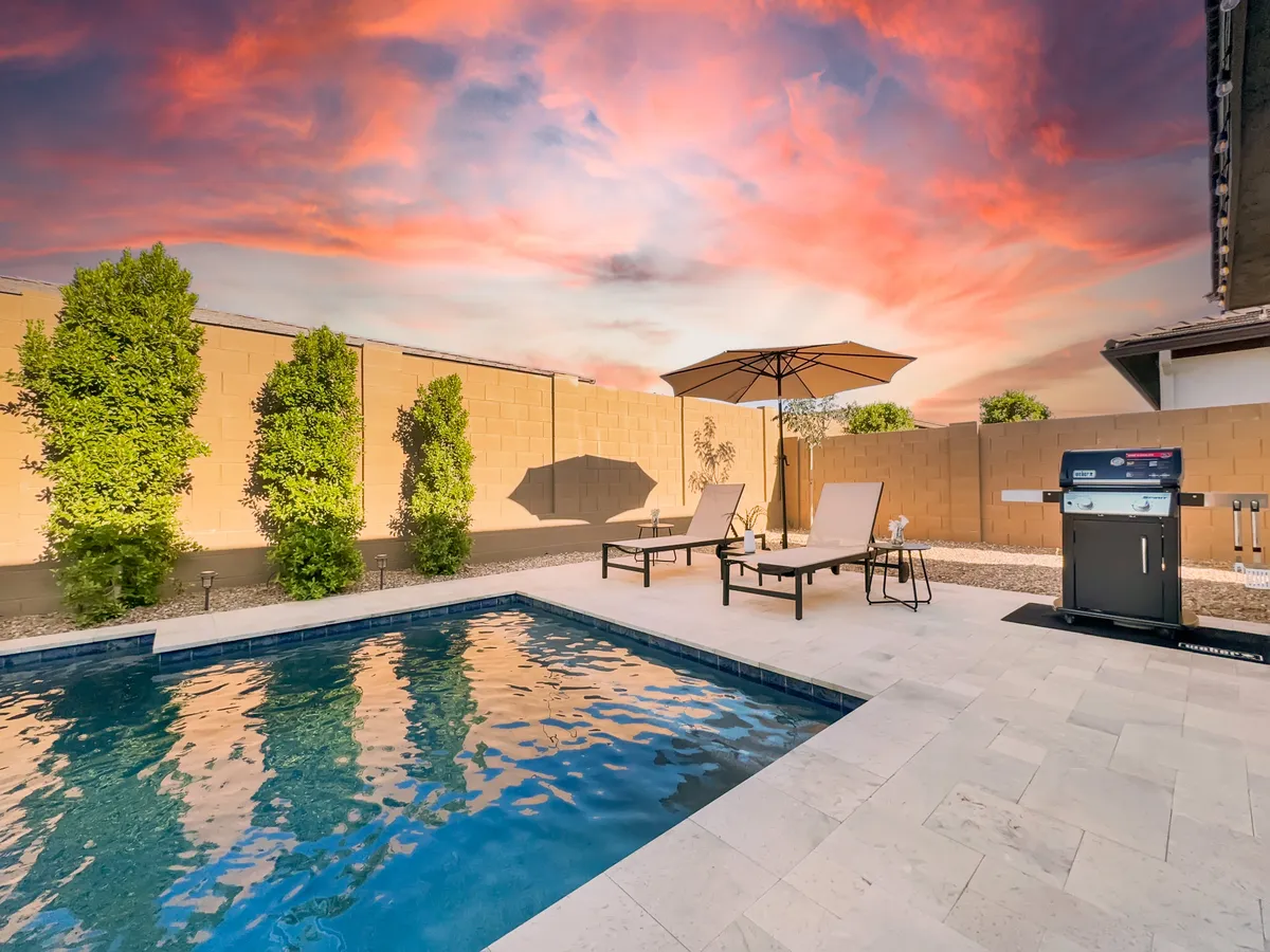 This exterior image captures the heart of the backyard oasis, showcasing a sparkling swimming pool, multiple lounge chairs, a BBQ grill, umbrella-shaded seating, and lush green shrubs. The setup is inviting and reflects spaces for swimming, sunbathing, grilling, and outdoor relaxation.