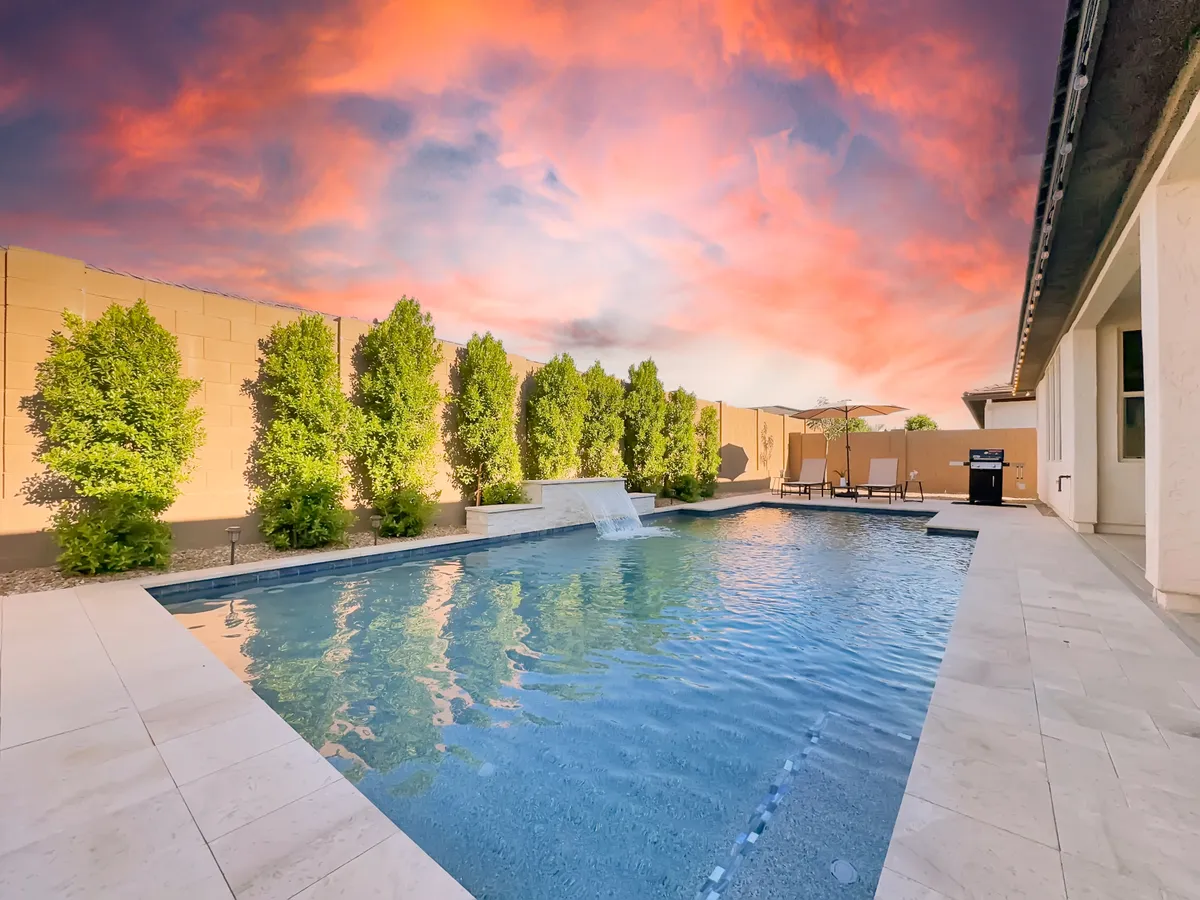 This image showcases a serene backyard featuring a sparkling swimming pool with a waterfall feature, lush green hedges, and inviting lounge chairs. The scene is set against a beautiful sunset sky, evoking a sense of relaxation and leisure in a private outdoor retreat.