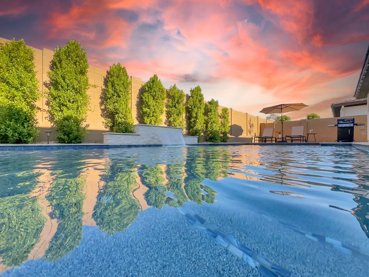 A serene exterior view of a swimming pool at sunset, with tall green trees and elegant patio furniture around the poolside. The tranquil atmosphere is ideal for evening gatherings, casual relaxation, or solitary moments by the water.