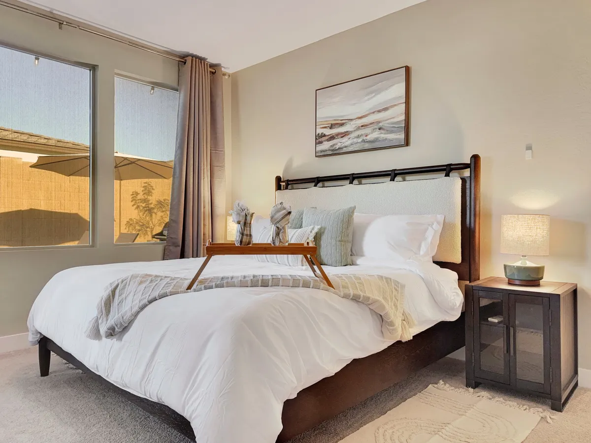 A tranquil bedroom with a bed dressed in decorative pillows, framed artwork above, a nightstand with a lamp and decor, and large windows letting in soft natural light. The setting is cozy and designed for relaxation.