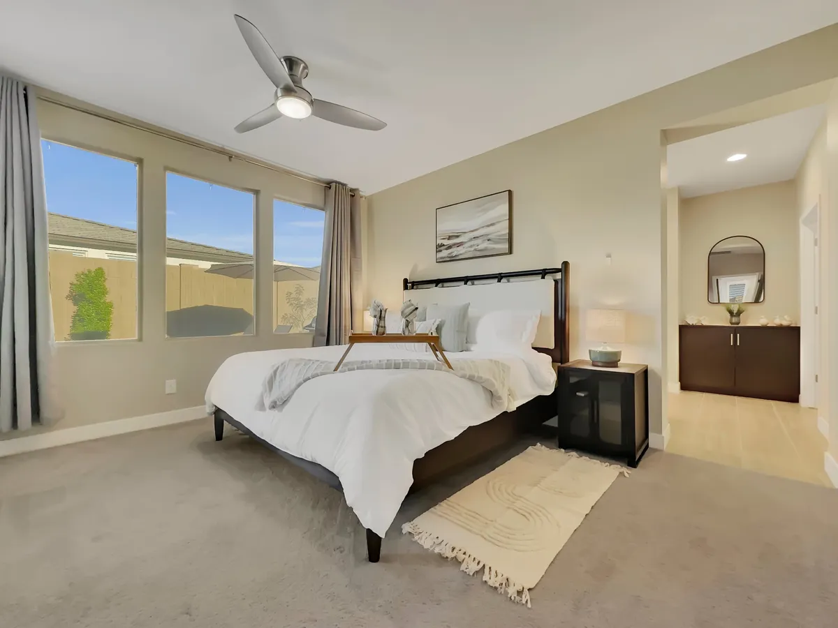 This image features a bedroom with a plush bed adorned in white bedding, flanked by nightstands with lamps and a decorative mirror. Large windows allow natural light to fill the space, creating a peaceful and relaxing atmosphere ideal for restful nights.