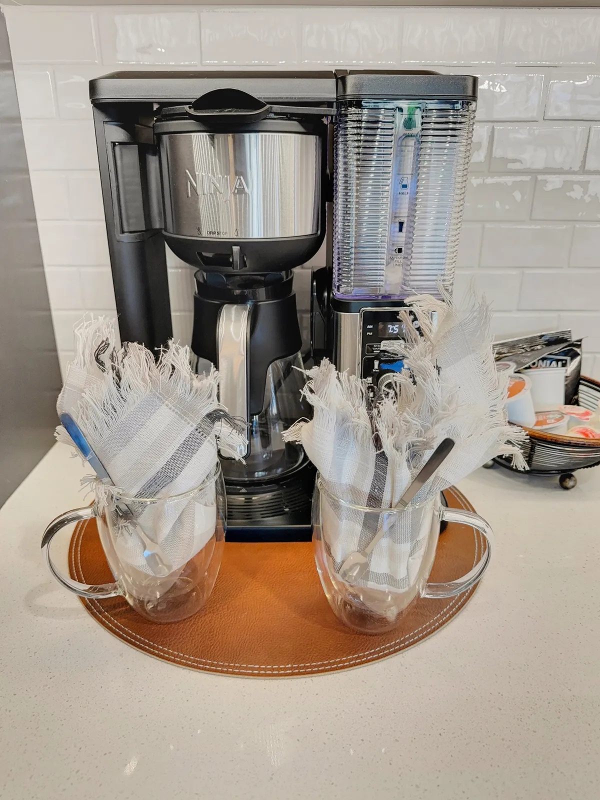 A close-up of a cozy coffee setup on a brown leather tray, featuring a Ninja coffee maker and two clear mugs ready for a morning (or midday) coffee break. The warm, inviting mood emphasizes comfort and daily rituals.